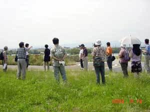 草が生えている淀川堤防上でワークショップの参加者たちが周囲に集まり、話しを聞いている写真