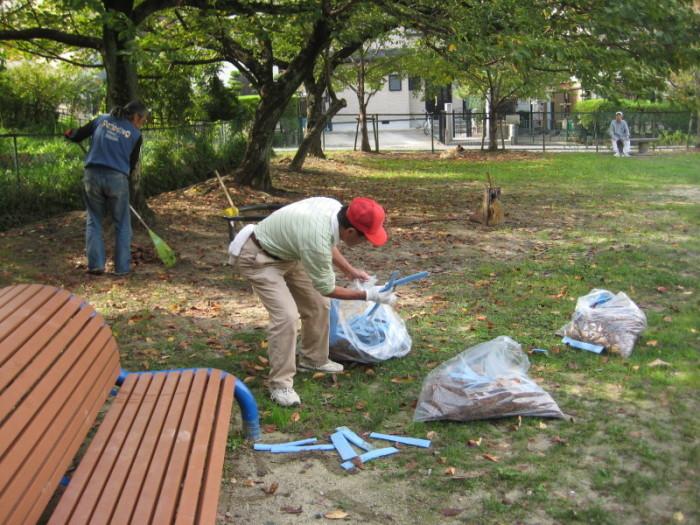 公園で落ち葉を熊手で集める青いベストを着用した男性とごみを袋に入れる赤い帽子を被った男性の写真