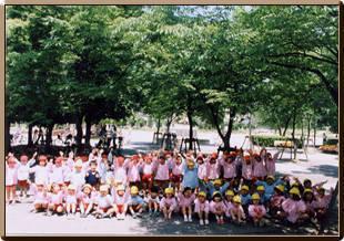 初本町公園の生い茂った樹木の前で前列の黄色い帽子を被った子どもたちが座り、後列には赤い帽子を被った子どもたちが立ち並び両手を広げて記念撮影している写真