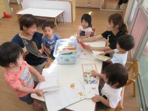 机の上に置かれた絵本を子供たちが座って眺めたり、お母さんに読み聞かせをしてもらっている写真