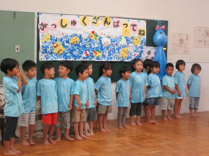 「がっしゅくがんばってね」と書かれた黒板の前に水色のTシャツ姿の子供たちが横一列に並んでいる写真