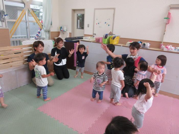 ピンクのマットの上や周りに子供たちが立っていて、保育士たちと一緒に手を上にあげたりしながら遊んでいる写真