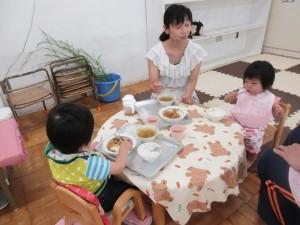 円型のテーブルに2人の赤ちゃんが座っていて、隣に座ったお母さんが食べる様子を見ている写真