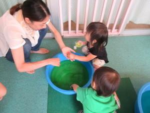 中央に置かれたたらいに緑色の水が入っていて、子供2人と女性が遊んでいる写真