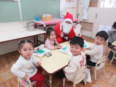 机に四人の子供が座っていて、奥に座っている子供の横に片手をあげたサンタクロースがしゃがんで並んでいる写真