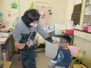 目隠しをした男の子が座っていて前に立っている女性が男の子の口に細長いお菓子のようなものをくわえさせている写真