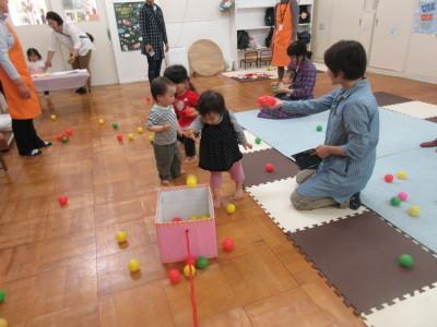 三人の子供がボールで遊んでいて、横に座っている一人のお母さんがボールを子供に渡そうとしている写真