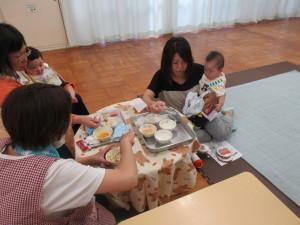 円形のテーブルに置かれた離乳食をお母さんが抱きかかえた赤ちゃんに食べさせようとしている写真