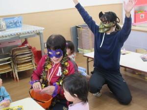 奥にお面をつけた保育士が座っていて、手前には紫のアイマスクをつけた保育士が子供たちにお菓子の様なものを配っている写真