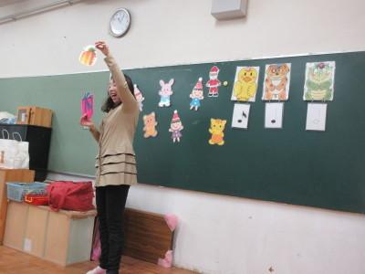 黒板の前に立った女性が右手にはプレゼントの絵、左手にはパイナップルの絵が描かれた紙を持って話をしている写真