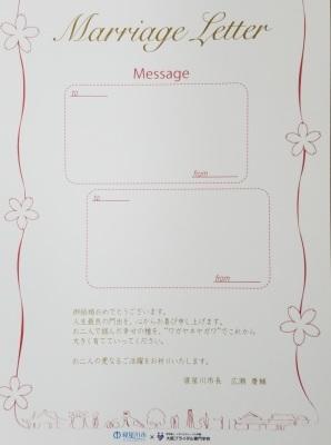 中央にメッセージを書く欄が2か所設けられ、両脇の赤い糸の上に花びらがあり、カード下部にまちの様子のシンプルなイラストと赤い糸が繋がってが描かれているメッセージカードの写真