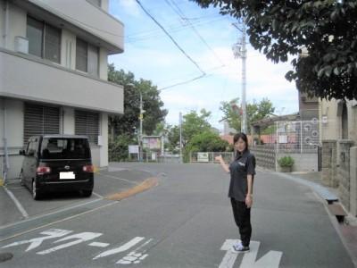 左側にアパートのような建物がある横の道路で、奥に見えているRELATTOの看板を指している女性の写真