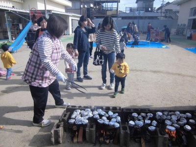 アルミホイルに包まれた焼き芋が網の上に並べられ、親子が焼き芋を貰いに来ている写真