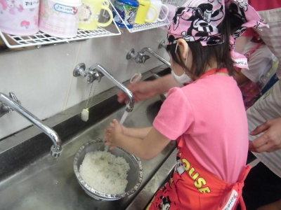 三角巾とエプロンをつけて手洗い場でざるに入った米を研いでいる子供の写真