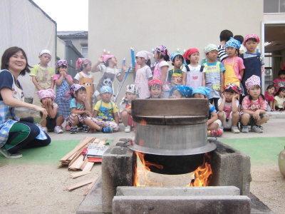 園庭に作った釜戸で大きな釜に入ったご飯を炊いている様子を周りで見ている子供たちの写真