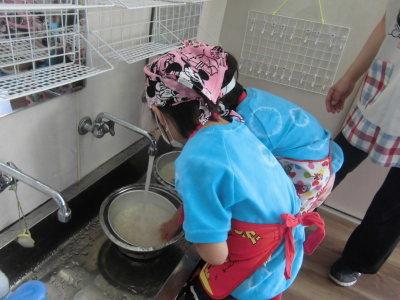 三角巾とエプロンをつけて、手洗い場でボウルの中の米を研いでいる2人の子供の写真