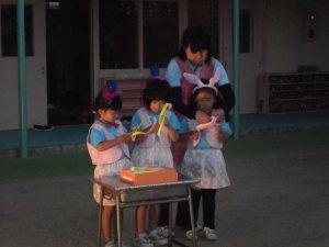 お揃いのベストとスカートを着て紙切りをしている3人の女の子とその後ろに立っている先生の写真
