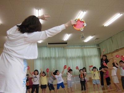 片手にタンバリンをもって、両手を頭の上に挙げて踊っている子供たちとお手本を見せている先生の写真