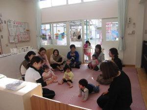 教室の床にまるく円を描くように座って絵本の読み聞かせを聞いている保護者と子供たちの写真