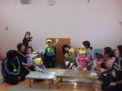 黄色い帽子をかぶりピンクや緑の衣装を着て保護者に抱っこされている子供たちの写真