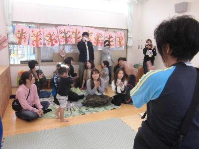 園児の絵の作品が飾ってある教室の床に座って手をたたいている保護者とその後ろからビデオカメラで撮影をしている保護者の写真