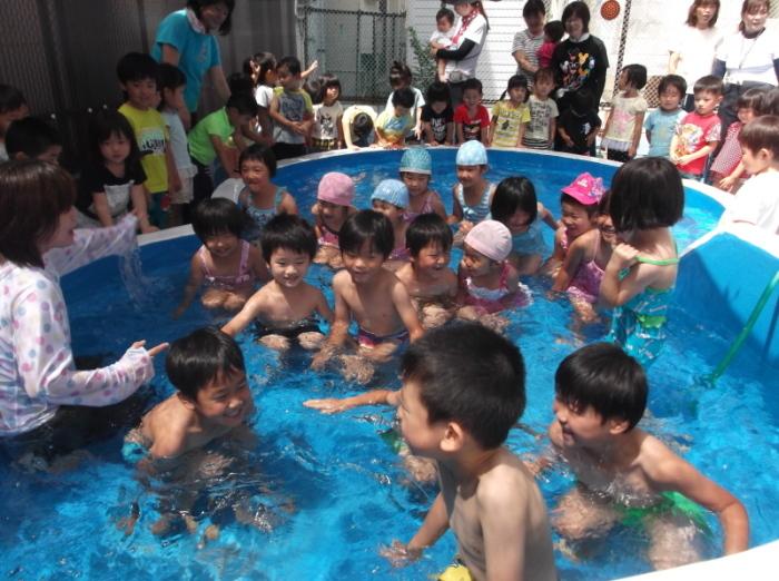 プールの中で肩まで水に浸かっている子供たちとプールの周りに集まってその様子を見ている子供たちと先生の写真