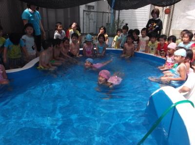 プールの中で泳いでいる水色とピンク色の水泳帽子をかぶった女の子と周りに集まって見ている子供たちと先生の写真