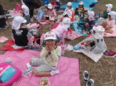 公園にレジャーシートを敷いてお弁当を食べている白い帽子の子供たちとカメラに向かってピースサインをしている女の子の写真
