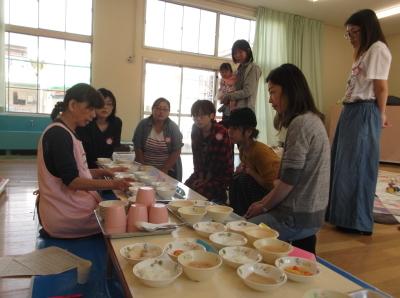 テーブルの上に並んだたくさんの種類の離乳食の見本と説明をしている給食の先生、熱心に話を聞いている母親の写真