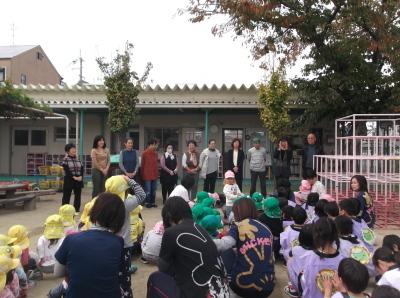 園庭に集まった子供たちと先生、前方に横一列に並んで立っている地域住民の方々の写真