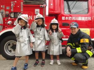 ヘルメットと消防服を着て消防車の前でポーズをきめている3人の女の子と消防士の写真