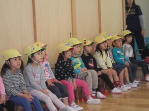 黄色い帽子をかぶり横一列に並んで椅子に腰かけている子供たちの写真