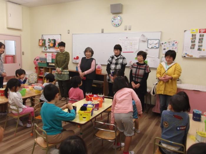 教室の前方に立っている5人の女性、毛糸やペットボトルが置かれた机にグループごとに分かれて席についている子供たちの写真