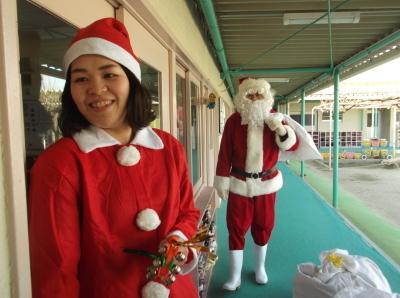 赤と白のクリスマスの衣装を着た先生とその後ろから歩いてくるサンタクロースの写真