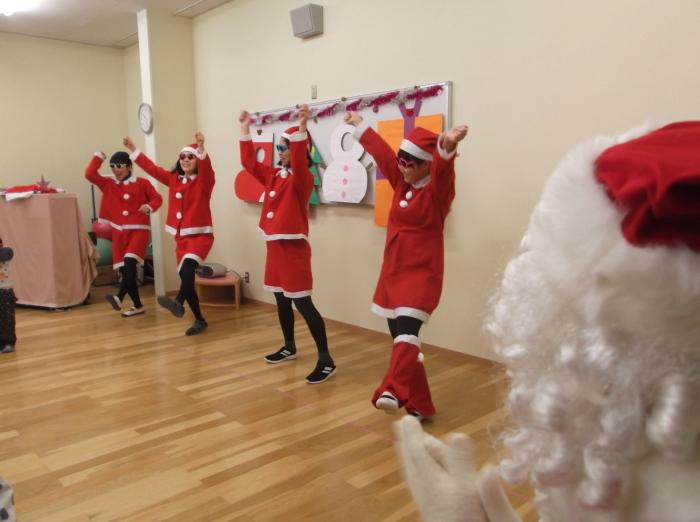 赤と白のクリスマスの衣装を着て踊っている4人の先生と横で手をたたいているサンタクロースの写真