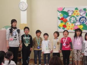 一番左に立っている先生、折り紙で作ったメダルを首から下げている子供たちが横一列に並んで立っている写真