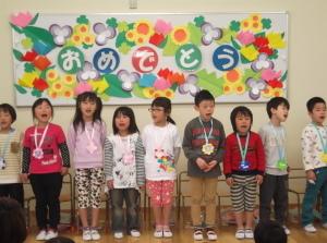折り紙や画用紙で作った花や「おめでとう」の文字が貼ってあるホワイトボードの前に立っている子供たちの写真