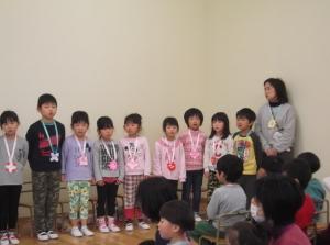 一番右側に立っている先生、その横に一列に並んで立っている子供たちと椅子に腰かけている子供たちの後ろ姿の写真