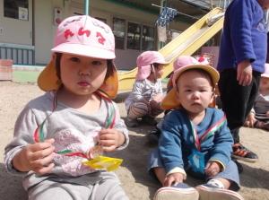 首から手作りのペンダントを下げて園庭に座っている2歳児の子供たちの写真