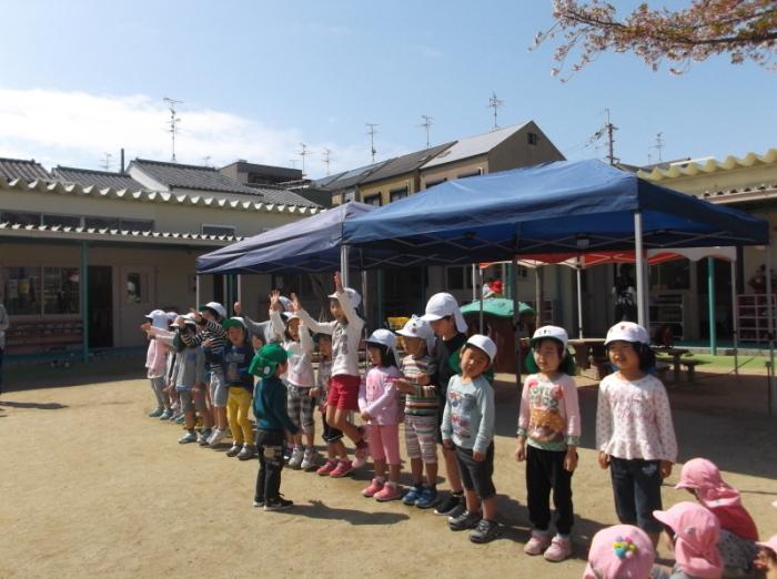 園庭に立てられた二張りのテントの前に横一列に並んで立っている白い帽子をかぶった子供たちの写真