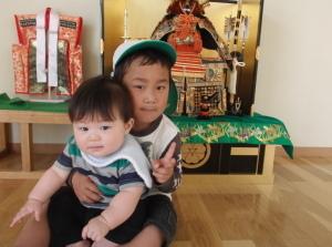 飾ってある武者人形の前でピースサインをしている男の子とその男の子に抱かれている赤ちゃんの写真