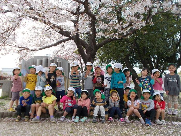 満開の桜の木の下に2列に並びピースサインや笑顔を浮かべる子ども達の写真