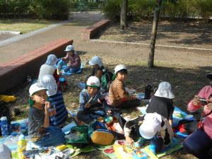 白い帽子を被った子ども達がシートを広げて友達と一緒に楽しそうにお弁当を食べている写真
