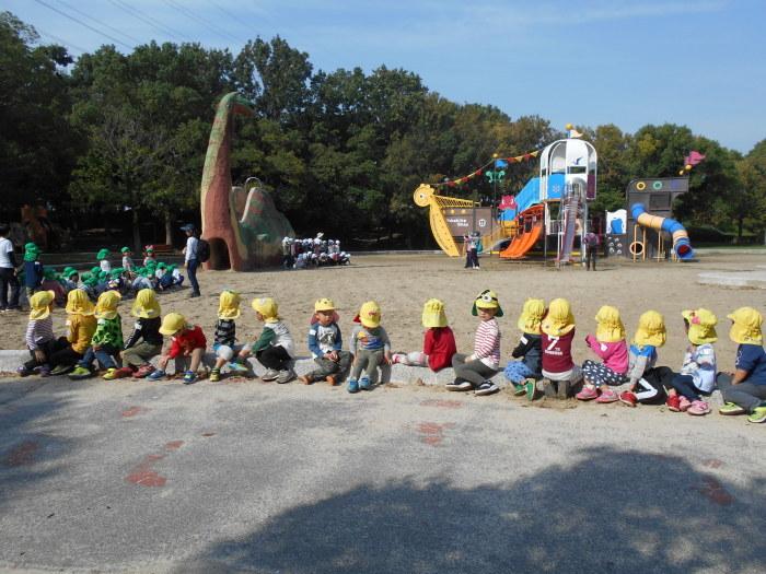 恐竜をモチーフにした遊具のある公園で、横一列に並んで座っている黄色い帽子を被った子ども達の写真