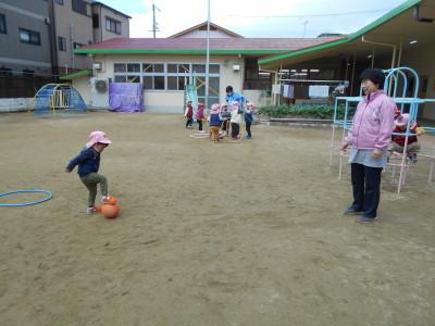 ピンク色の帽子を被った男の子が足でボールを止めており、その様子を少し離れた場所から先生が見守っている写真