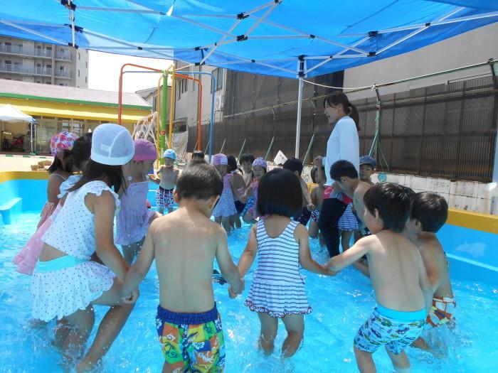 ビニールシートの日よけのあるプールで、水着を着た子ども達と先生が手をつないで大きな円を作って足踏みしている写真