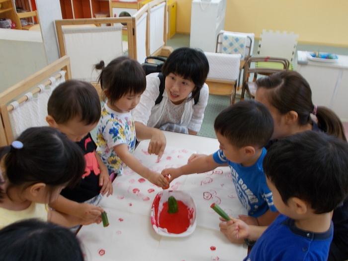 机の上に置かれた赤い絵の具にきゅうりをいれスタンプ遊びをしている園児達の写真