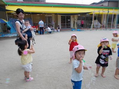 園庭でシャボン玉遊びをしている園児達をそれを見守る1組の親子の写真