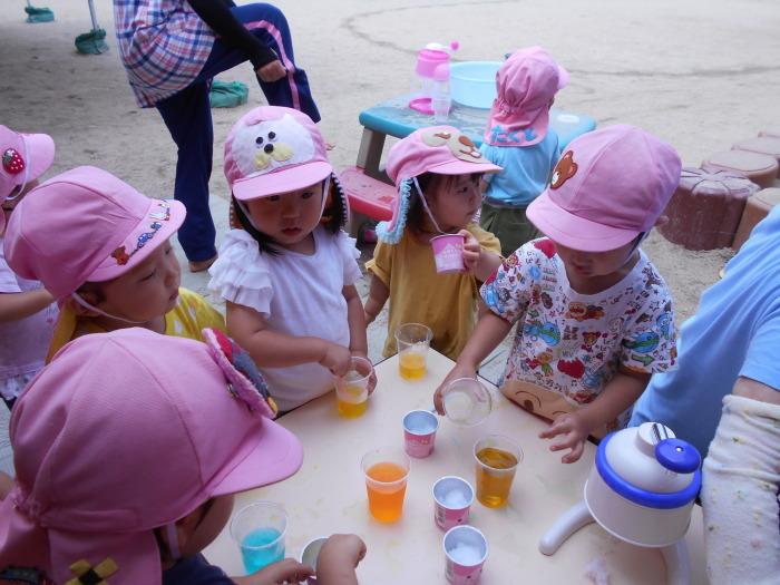 ピンク色の帽子を被った子どもたちが机の上に置かれた水色や黄色の液体が入ったコップや氷を触って遊んでいる写真