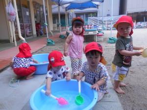 水色のたらいの中に入っているお水をスコップですくって遊んでいる赤い帽子を被った子ども達の写真